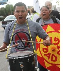 Latino man in foreground with drum around his neck and hitting it with drumsticks, his mouth open chanting, wearing a shirt that says Boycott Wendys with a picture of a girl's head with red pigtails and a No sign around it (circle with line through it) and a Latino woman behind him holding a bright orange and yellow sign also with her mouth open chanting