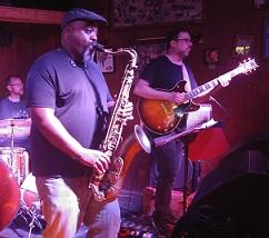 Heavy set black man in a cap playing the saxophone with other guys in a band onstage