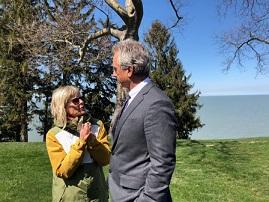 Short blonde wtoman wearing a yellow coat and sunglasses talking and gesturing to a taller white man with gray hair and a gray suit outside by tree and a lake 
