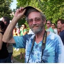 White man raising up his cap to the camera, smiling with sunglasses and gray mustache and beard, wearing a blue tye-dye t-shirt standing outside around trees and a lot of other people 