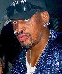 Black man in a baseball cap with lots of facial rings in lips, nose and ears wearing a blue shirt