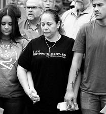 Black and white photo of three Latino people, a young girl with long brown hair looking down, a middle-aged woman with a black T-shirt and necklace looking down and a young man looking very worried, all holding hands with several people behind them 