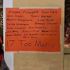 A sign on orange paper taped to a brown wall listing all the names of people killed in the recent Parkland Fla school shooting and the words 17 too many at the bottom 
