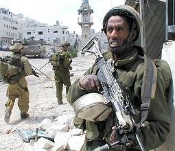 Foreground has a dark skinned man holding as assault rifle in the background two military men with rifles walking toward a building