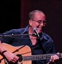 Balding white man with glasses wearing a blue button down shirt singing into a mic and playing a guitar