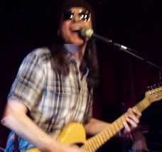 Man with long brown hair in plaid shirt playing a guitar and singing into a mic