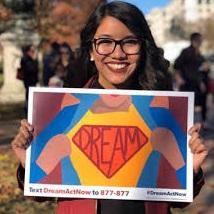 Young smiling woman with long black hair and big black-rimmed glasses holding a bright colorful sign that says DREAMER on it