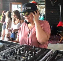 Woman with a baseball cap on and headphones at a mixing board smiling, with people in the background
