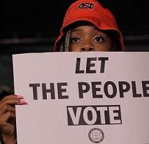 Black person's face with a red hat on their head peering over a sign that they are holding that says Let The People Vote