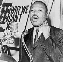 Black man speaking at a mic wearing a suit, with a sign in the background to his left saying Why We Can't