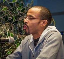 Young black man facing left talking into a microphone, he has short cropped hair and glasses and a goatee, sitting in front of a plant