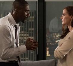 On the left a black man facing right in a white shirt and tie looking at a woman his hands in front of him with fingers clasped like he's praying and the white woman with long brown hair to the left is looking up at him with arms folded wearing a pick shirt. They are standing in front of a window looking over a nighttime city scene