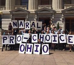 Big white capitol building with columns and turrets above windows in the background, lots of people posing in front holding placards each with a letter spelling out NARAL PRO-CHOICE OHIO