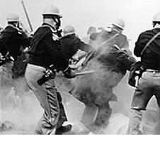 Black and white photo of police wearing helmets with their backs to the camera, looking like the are beating people with sticks among a lot of gas in the air obscuring the people in the picture