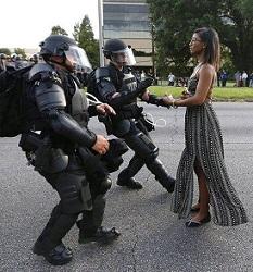 Young black woman in long flowing black and white dress wearing glasses standing straight and tall and calm and two older white heavily armed riot police looking like they are stepping back away from her