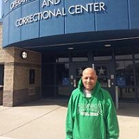 Bald older man wearing a green pullover sweatshirt that says Capital University standing in front of a building with glass doors and a glue curvy sign above that reads ...and correctional center