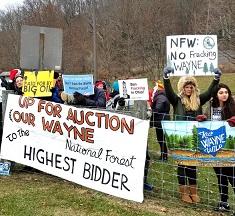 Lots of young people wearing coats outside near a forest at a fence with protest signs reading Up for Auction Our Wayne to the Highest Bidder