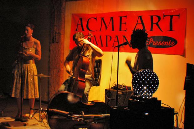 A red sign sayigng ACME ART on the wall behind silhouettes of musicians, a guy playing a bass fiddle, a person singing into a mic and a woman wearing a mid-length dress