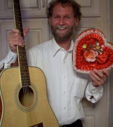 Man with a beard and a big smile and wearing a white button down shirt standing in front of a door holding a guitar and a red heart shaped box