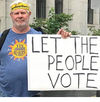 White man with yellow headband and blue T-Shirt with sun in the middle holding a sign that says Let the People Vote