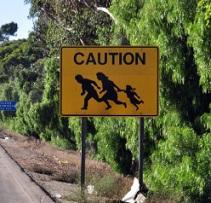 Yellow sign by the road and a hill with silhouettes of a family holding hands and running and the words CAUTION