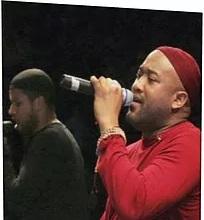 Black men in concert, one in red singing into mic