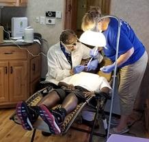 Person in a dental chair and a dentist to the left working on them and a woman to the right, both with mask and gloves on
