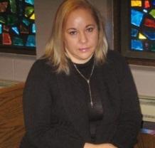 Young Latina woman with blonde hair and black clothes sitting on a pew in a church with stained glass windows behind her