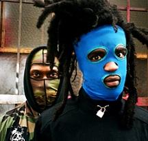Two men one in front with dreds sticking up off his head and a bright blue mask and a guy behind him with a camouflage mask