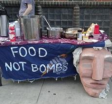 Big paper mache face hanging on the front of a table outside next to a blue sign with white hand drawn letters that say Food Not Bombs and a lot of pots and pans and condiments on the table and a brick wall in the background