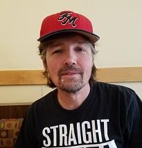 Man with brown hair sticking out from a red baseball cap and goatee wearing a black T-shirt 