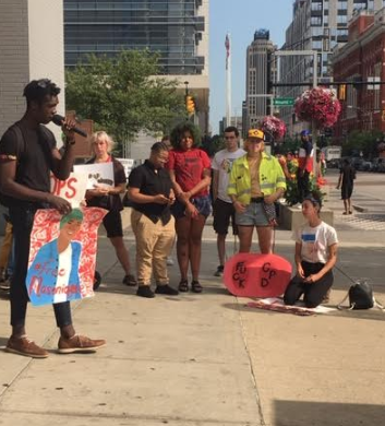 Young black man speaking into a mic to a crowd of people outside with signs