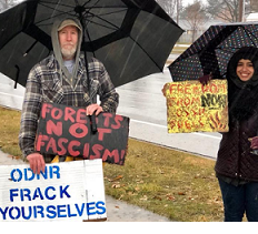 Two people standing outside in the rain holding umbrellas and signs, one says ODNR Frack Yourselves