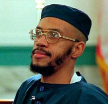 Black man with wire rimmed glasses and mustache and beard and a square topped blue hat