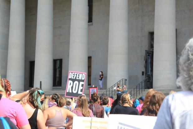 crowd protesting in front of state house with sign that says defend roe