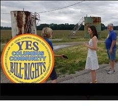 People standing outside in front of weird looking rusty metal structures and the round yellow logo for Columbus Community Bill of Rights