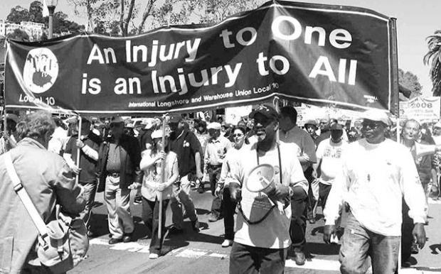 Black and white photo of big banner with words An Injury to one is an injury to all and mostly black people marching with it, some with drums