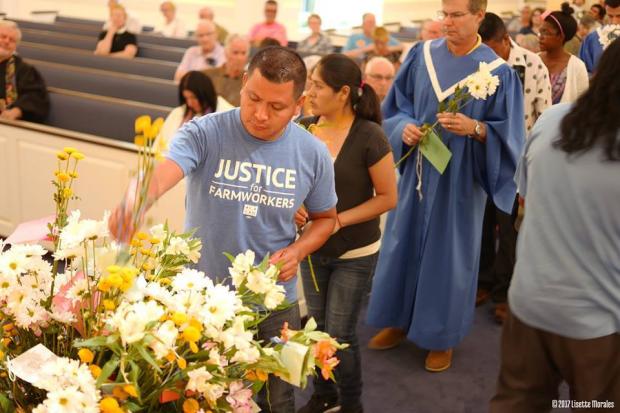 Main in a Justice for Farmworkers light blue T-shirt putting flowers into a huge container that has a lot of flowers in it and a line of people behind him and pews of a church behind them
