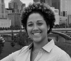 Smiling black woman with short cropped curly hair and a striped button down shirt standing with downtown buildings and a bridge across the river behind her