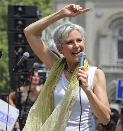 Jill Stein with her arm above her head and in a white tank top and yellow scarf outside at rally