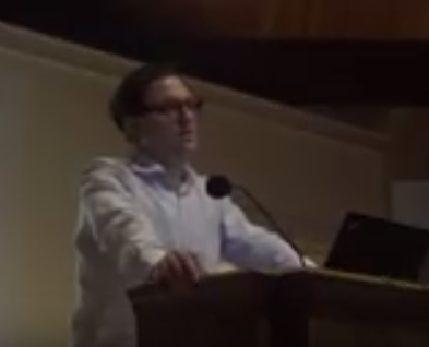 White man with glasses and white shirt standing and speaking at podium