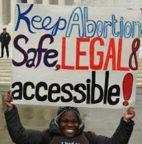 Black woman holding sign saying Keep abortion safe, legal and accessible in from of a government building with a cop standing in the background