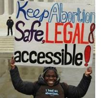 Young black woman smiling with winter clothes on outside an official looking government building holding a sign above her head saying Keep Abortion Safe, Legal & accessible! A cop is standing back in the distance.
