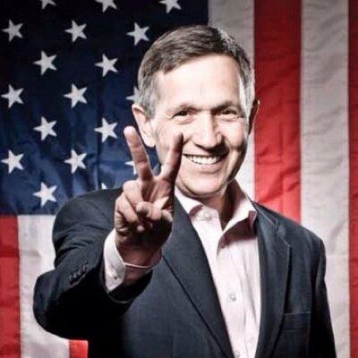 White middle aged man smiling an making a peace sign in front of a flag
