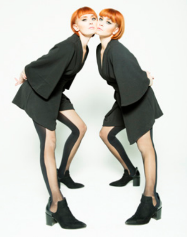 Two women with red hair and black outfits leaning towards each other