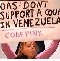 Blonde woman in pink shirt holding big sign above her head that says OAS: Don'ts support a coup in Venezuela