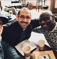 A balding white guy with red framed glasses and facial hair smiling sitting at a table with his arm around a bald black man in a white and black designed sweater also smiling