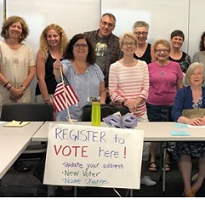People posing standing behind a white table with a sign that says register to vote here