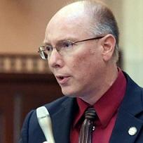 Balding white guy with sire rimmed glasses, a blue suit coat and maroon shirt and striped tie holding a silver microphone talking into it