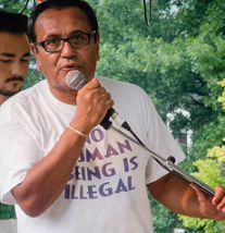 Latino man in T-shirt that says No Human Being is Illegal, talking into a mic